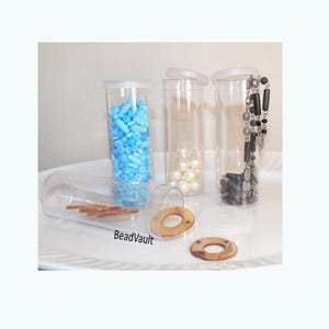 May include: Four clear cylindrical plastic storage containers with white lids. One container is filled with light blue beads, one with white pearls, one with a black and silver necklace, and one lying on its side contains brown ring-shaped beads.