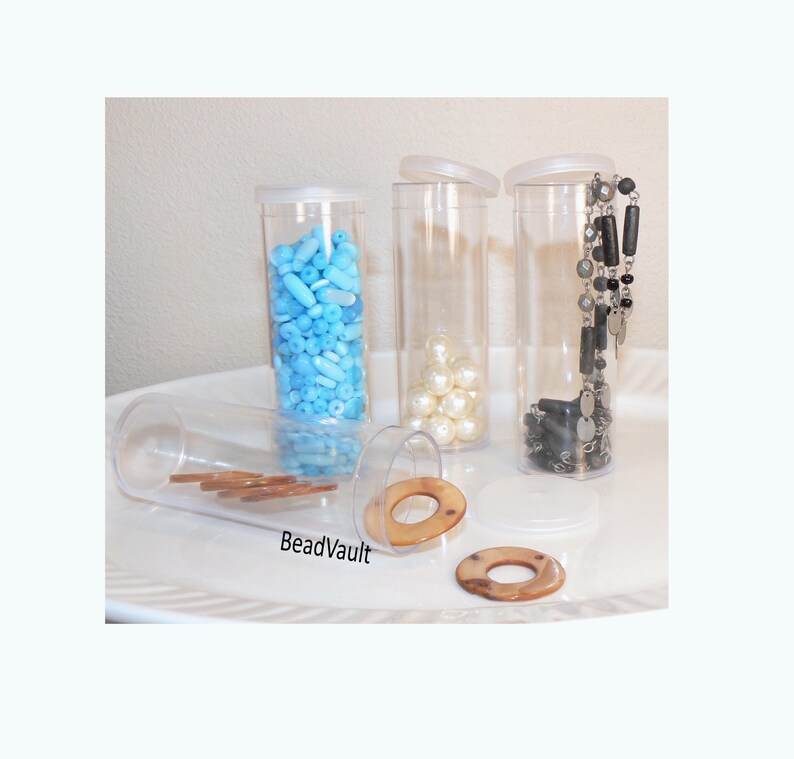 May include: Four clear cylindrical plastic storage containers with white lids. One container is filled with light blue beads, one with white pearls, one with a black and silver necklace, and one lying on its side contains brown ring-shaped beads.