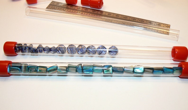 May include: Two clear plastic tubes with red caps filled with blue and green beads. The tubes are on a white surface.