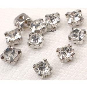 May include: Ten clear crystal rhinestones with silver settings. The rhinestones are square shaped and have a faceted design. The rhinestones are arranged in a scattered pattern on a white surface.