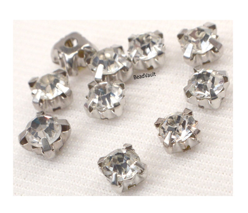 May include: Ten clear crystal rhinestones with silver settings. The rhinestones are square shaped and have a faceted design. The rhinestones are arranged in a scattered pattern on a white surface.