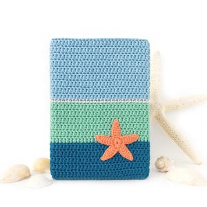 May include: A rectangular crochet cover featuring horizontal stripes in light blue, mint green, and dark blue. A textured orange crochet starfish is attached to the lower section. The item is surrounded by several small white seashells.