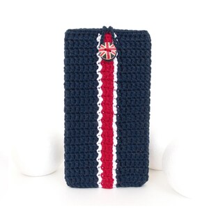 Puede incluir: Una funda para teléfono de crochet azul marino con una franja roja, blanca y azul en el centro. La funda tiene un botón con un diseño de Union Jack.