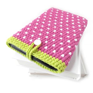 May include: A pink crocheted tablet case with white heart-shaped accents. The case has a lime green border and a white button closure. The case is resting on a stack of white books.