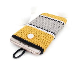 May include: Crocheted phone case in yellow, gray, black, and white. The case has a button closure and a black loop. The phone is partially visible inside the case. The case is made of yarn.
