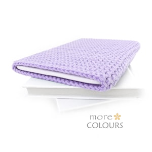 May include: A lavender crocheted laptop sleeve with a white lining. The sleeve is folded over a stack of white books. The text "more colours" is printed on the bottom of the image.