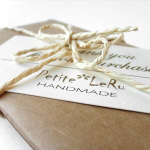 Puede incluir: Un paquete de papel marr&oacute;n envuelto con una cuerda de rafia de color beige claro atada en un lazo. Una tarjeta rectangular blanca est&aacute; metida bajo la cuerda, con el texto impreso "Thank you for your purchase!" y la marca "Petite LeRu HANDMADE".