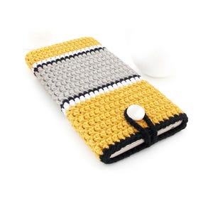 May include: A handmade crochet phone case in yellow, gray, black, and white. The case features a button closure and a striped design. The case is designed to fit a smartphone.