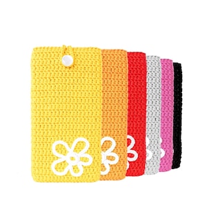 Op de afbeelding: Zes gehaakte telefoonhoesjes in verschillende kleuren, waaronder geel, oranje, rood, wit, roze en zwart. Elk hoesje heeft een wit gehaakt bloemetjesontwerp.