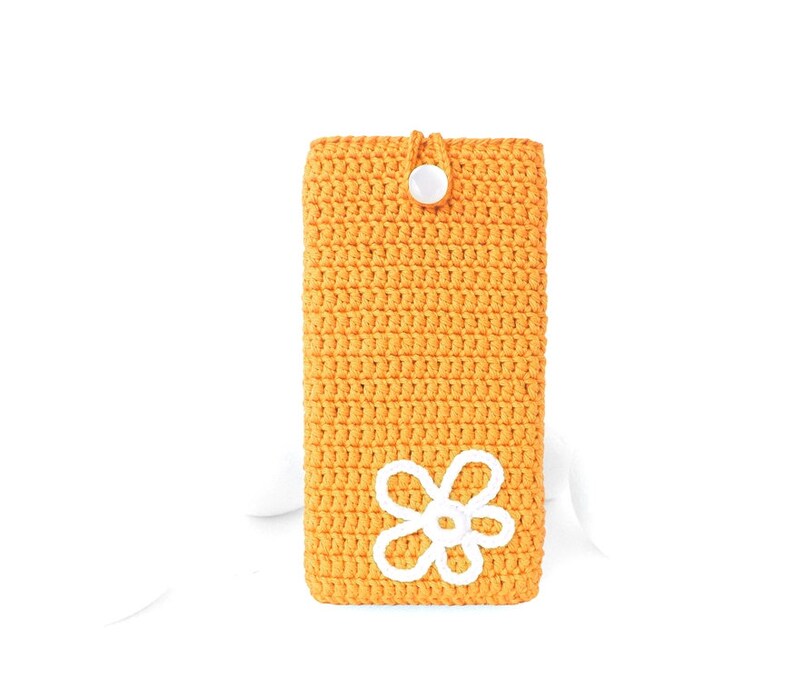 Op de afbeelding: Een gele gehaakte telefoonhoes met een wit gehaakt bloemenmotief en een witte knoopsluiting.