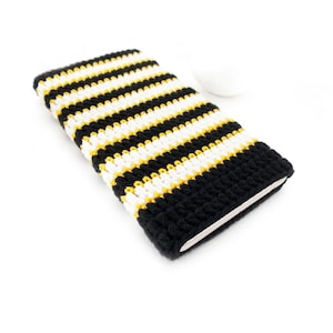 Puede incluir: Funda de teléfono tejida a ganchillo con un diseño de rayas negras, blancas y amarillas. La funda rectangular está diseñada para un smartphone. El tejido de ganchillo apretado sugiere un artículo hecho a mano.