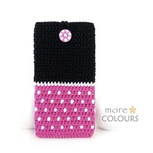 Puede incluir: Funda de teléfono de ganchillo hecha a mano en negro y rosa. La parte superior es negra y la inferior rosa con lunares blancos. Un borde blanco separa los colores. Un botón rosa y blanco asegura la solapa superior. El texto "more COLOURS" está en la esquina inferior derecha.