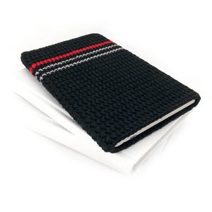 May include: A rectangular black crochet sleeve featuring a textured knit pattern with a single horizontal red stripe and a thin white stripe near the top edge. The sleeve is designed to hold a flat electronic device or notebook securely inside.