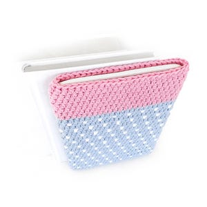 May include: A handmade crochet book cover in pink and light blue. The top half is solid pink, while the bottom half features a light blue dotted pattern. The cover is designed to hold a book, with the pages visible.