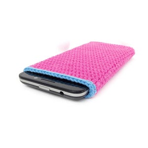 May include: A rectangular smartphone case made of bright pink crocheted yarn with a light blue trim along the opening. The case is shown holding a black smartphone with a visible camera lens and a headphone jack on the top edge of the device.