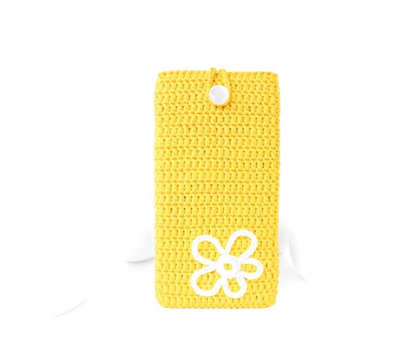 Op de afbeelding: Een gele gehaakte telefoonhoes met een wit gehaakt bloemenmotief en een witte knoopsluiting.