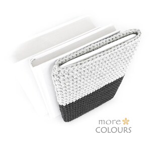 May include: A crocheted book cover in gray and black, with the text "more COLOURS" in the bottom right corner. The cover is wrapped around a stack of white books, creating a stylish and protective accessory.