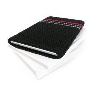 May include: Black crochet tablet sleeve with a red and gray stripe detail. The sleeve is resting on a stack of white paper. The tablet is visible inside the sleeve.