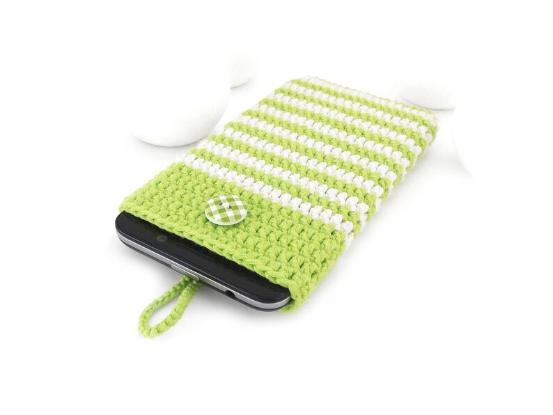 Puede incluir: Una funda de ganchillo para tel&eacute;fono con rayas horizontales de color verde lima y blanco. La funda tiene un peque&ntilde;o lazo verde lima en la parte inferior y se asegura con un bot&oacute;n redondo de cuadros verdes y blancos, conteniendo un m&oacute;vil negro.