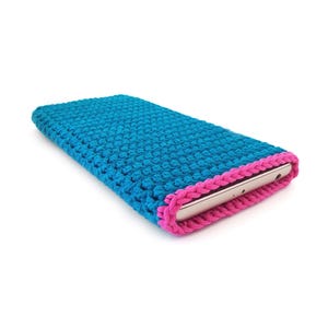 Puede incluir: Una funda de teléfono de ganchillo azul turquesa con un borde rosa. La funda es rectangular y parece hecha a mano. El teléfono es parcialmente visible en el interior.