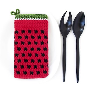 May include: A red and black crocheted watermelon-shaped hot pad with a green top, next to a white plate and two black serving utensils.
