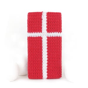 May include: A rectangular crochet cover featuring a vibrant red background with a prominent white cross pattern centered vertically and horizontally. The textile has a consistent, textured stitch throughout, creating a structured and uniform appearance.
