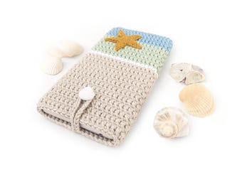 Puede incluir: Una funda de teléfono de ganchillo rectangular con una base beige, una sección central verde claro y una sección superior azul claro. Una estrella de mar de ganchillo color mostaza está fijada arriba y un cierre de botón blanco asegura la solapa.