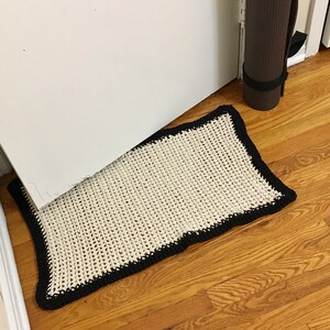 May include: A white crocheted doormat with a black border. The doormat is lying on a wooden floor.