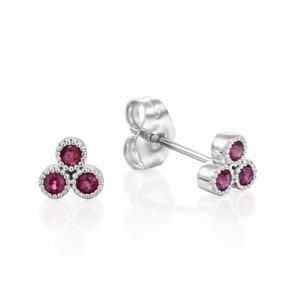 May include: A pair of silver stud earrings, each featuring three round, deep red gemstones. The gemstones are set within a beaded border, creating a cluster design. The earrings are displayed on a white background.