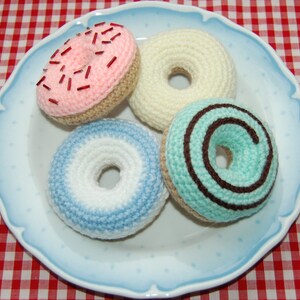 Op de afbeelding: Vier gehaakte donuts op een blauw-wit bord. De donuts zijn roze met rode sprinkles, wit, lichtblauw met witte strepen en lichtgroen met een bruine swirl.