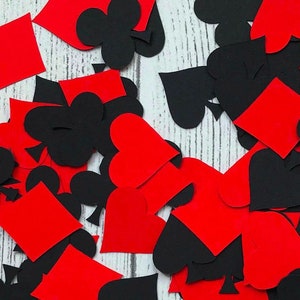 Puede incluir: Recortes de papel rojo y negro de palos de cartas de juego, incluyendo corazones, diamantes, picas y tr&eacute;boles. Los recortes est&aacute;n esparcidos sobre una superficie de madera blanca.