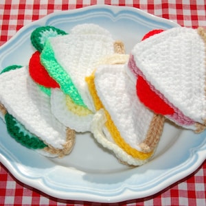 May include: Four crocheted sandwiches on a blue and white plate. The sandwiches are made with white yarn and have different coloured fillings, including green, red, and yellow.