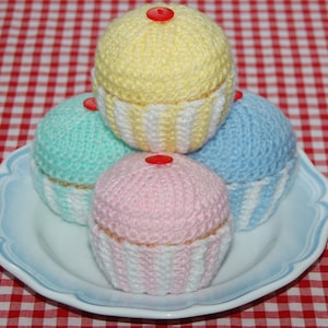 May include: Four knitted cupcakes in a stack on a white plate with a red and white checkered tablecloth. The cupcakes are yellow, light blue, pink, and green. Each cupcake has a red button on top.