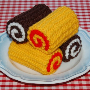 May include: Three crocheted rolls on a white plate with a blue rim. The rolls are yellow with red and white details. The rolls are on a red and white checkered tablecloth.