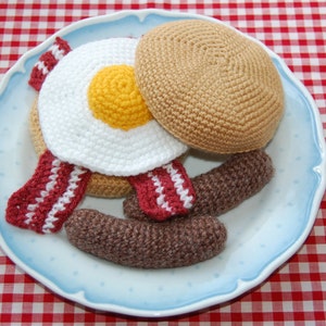 May include: A crocheted breakfast set on a blue and white plate. The set includes a brown pancake, a white egg with a yellow yolk, and two brown sausage links. The set is surrounded by red and white crocheted bacon.