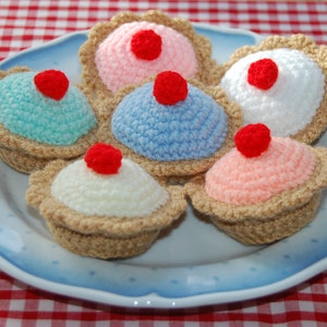 May include: Six crocheted cupcakes on a blue and white patterned plate. The cupcakes are decorated with red, pink, blue, and white frosting and red cherries.