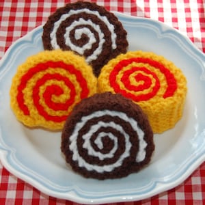 May include: Three crocheted swirl cakes on a white plate with a blue rim. The cakes are yellow, red, and brown with white swirls.