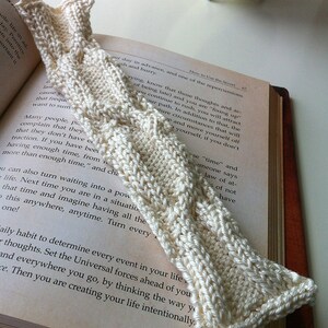 Puede incluir: Un marcapáginas de punto blanco con un patrón texturizado se coloca sobre un libro abierto con el texto "How to Use the Secret" visible. El libro está abierto en una página con texto sobre el tiempo y cómo usarlo a tu favor.