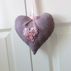 Könnte beinhalten: Ein lavendelfarbenes, gestricktes Herzförmiges Kissen mit einem rosa Band und einem herzförmigen Muster aus rosa Knöpfen.