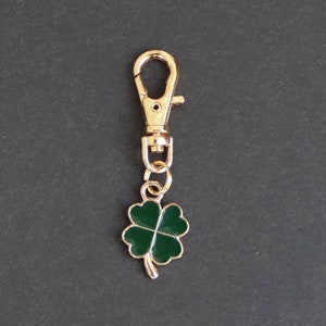 May include: Gold-colored keychain clip with a green four-leaf clover charm. The clover has a gold outline and a dark green enamel fill. The clip is a lobster claw style, and the charm is attached with a small ring.