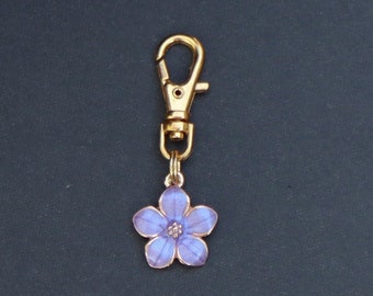 May include: A gold-colored keychain with a purple flower charm. The flower has five petals and a yellow center.