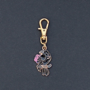May include: A gold-toned keychain featuring a black raven with a pink rose in its beak. The raven is perched on a branch and has a star-shaped design on its chest.