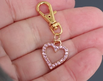 Könnte beinhalten: Ein goldfarbener Metallherz-Anhänger mit rosa Strasssteinen. Der Anhänger ist an einem goldfarbenen Karabinerverschluss aus Metall befestigt. Der Anhänger wird in der Hand einer Person gehalten.