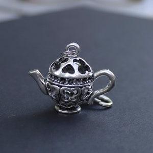 May include: Silver charm in the shape of a teapot with a delicate, intricate design. The teapot has a heart-shaped lid and a handle with a loop at the end.