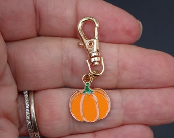 May include: A gold-colored metal pumpkin charm with an orange enamel finish and green stem. The charm is attached to a gold-colored lobster clasp. The charm is a fall-themed accessory.