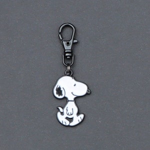 May include: A Snoopy keychain charm in a sitting pose, featuring a white body with black accents on the ears and head. The charm is attached to a metal clasp, perfect for keys or bags. The background is a solid gray.