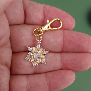 May include: A gold-toned metal lobster clasp charm featuring a snowflake-shaped pendant. The pendant is encrusted with multiple clear, marquise-cut and round-cut cubic zirconia stones arranged in a symmetrical, multi-layered floral star pattern.