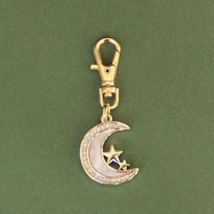 May include: A gold-colored keychain charm featuring a crescent moon and two stars. The moon has a white center and is outlined with small, clear crystals. The charm is attached to a gold-colored clasp.