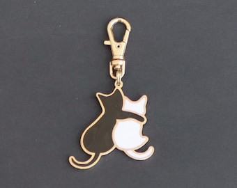 May include: A gold-colored keychain featuring two cats hugging. The cat on the left is black and the cat on the right is white.