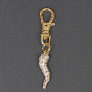 May include: Gold-toned metal charm with a lobster clasp and a small, curved horn pendant. The horn is encrusted with small, sparkling crystals. The charm is designed to be attached to a keychain or bag.